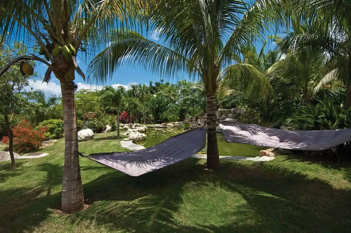 Grey cushioned hammocks hang between coconut palms over a green lawn, where a stone-ringed pond and red-orange foliage create a tropical garden retreat.