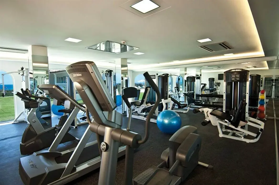 Professional cardio machines and weight equipment fill a bright gym with floor-to-ceiling windows, where mirrors reflect an ocean view beyond a green lawn.