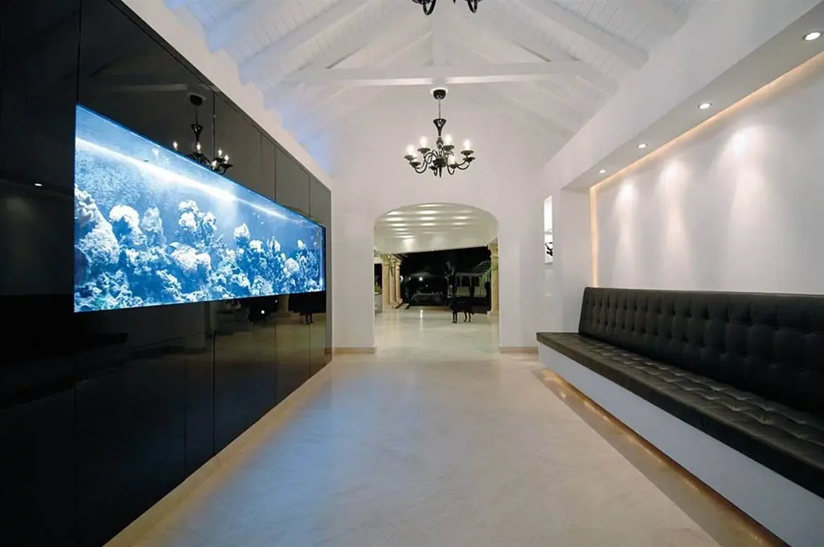 A glossy black wall with a glowing saltwater aquarium spans a vaulted hallway, where a tufted black leather bench and a black chandelier lead to an arched doorway.