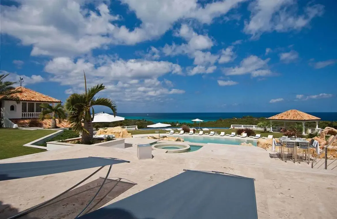 A circular hot tub cascades into a turquoise pool beside a terracotta-roofed dining terrace, where white loungers and green palms overlook a deep blue ocean.