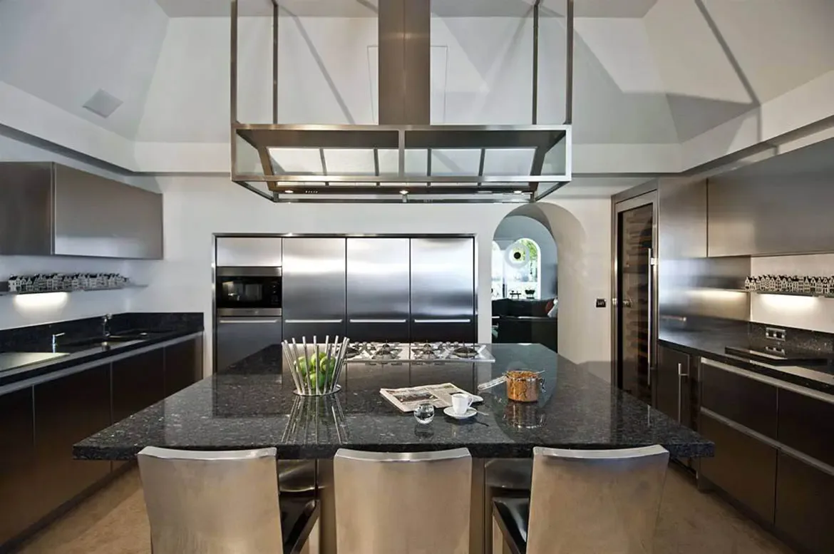 A dark granite island with metal stools sits under a geometric steel range hood in a vaulted white kitchen, where glass cabinetry and a wine fridge gleam beside an arched doorway.