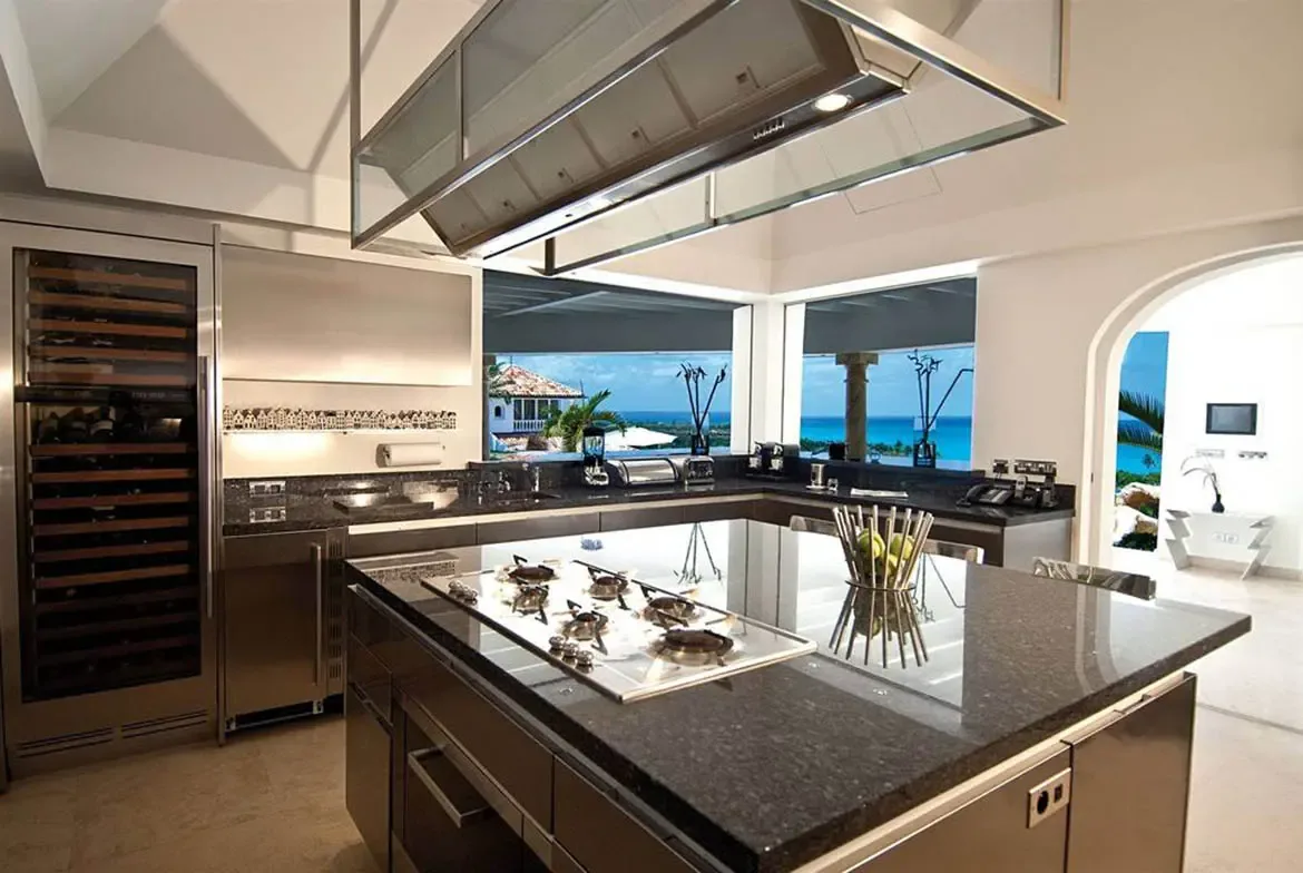 A stainless steel range hood and dark granite island anchor a professional kitchen with a wine fridge, where windows frame a turquoise ocean beyond palms.