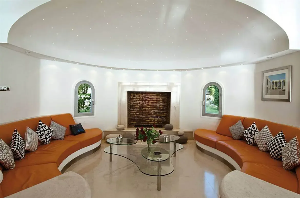 Two curved orange leather sofas circle a glass table with red roses in a domed room with a starry ceiling, where arched windows reveal lush greenery beyond a brick fireplace.