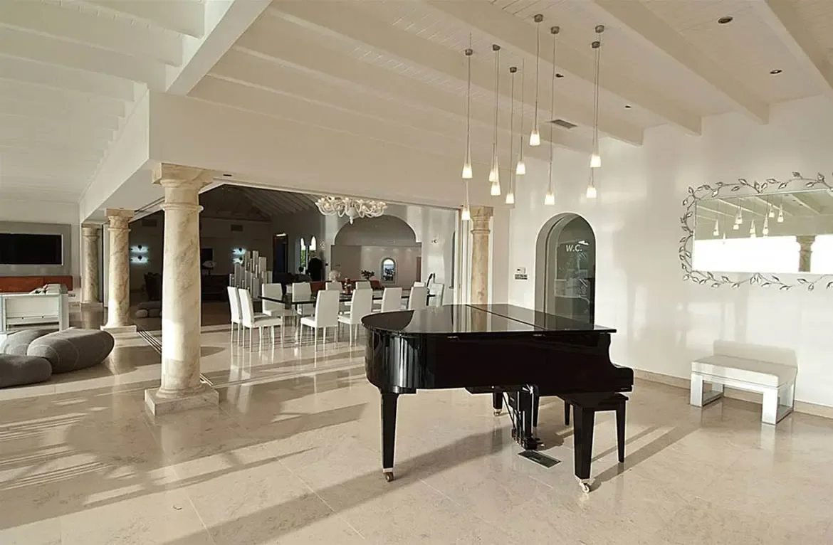 A black grand piano sits beneath clustered pendant lights on a polished marble floor, where travertine columns frame a glass dining table and an ornate mirror near an arched doorway.