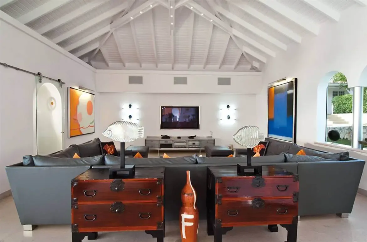 A grey L-shaped sofa with orange pillows faces a flat-screen TV under a vaulted ceiling, where abstract art and translucent fish sculptures flank an entertainment console.