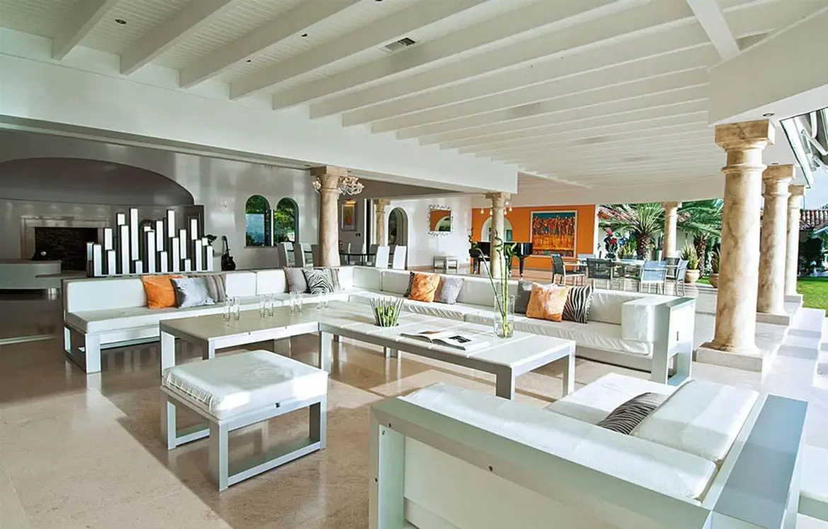 White sectionals with zebra-print and orange pillows cluster around glass coffee tables under a vaulted ceiling, where a skyscraper-light sculpture and a grand piano anchor the space.
