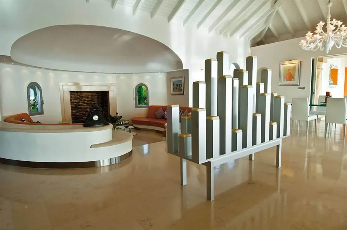 A sunken circular pit with orange cushions and a fireplace sits beside a skyscraper-like silver sculpture, where a branching chandelier hangs over a glass dining table.