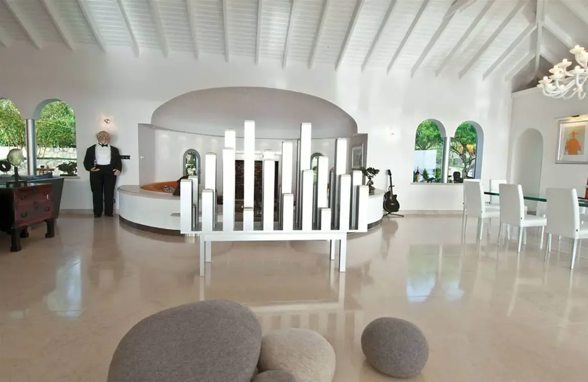 Grey pebble-shaped floor cushions and a white coffee table sit under classical stone pillars, where a grand piano and an orange accent wall with abstract art define the vaulted space.