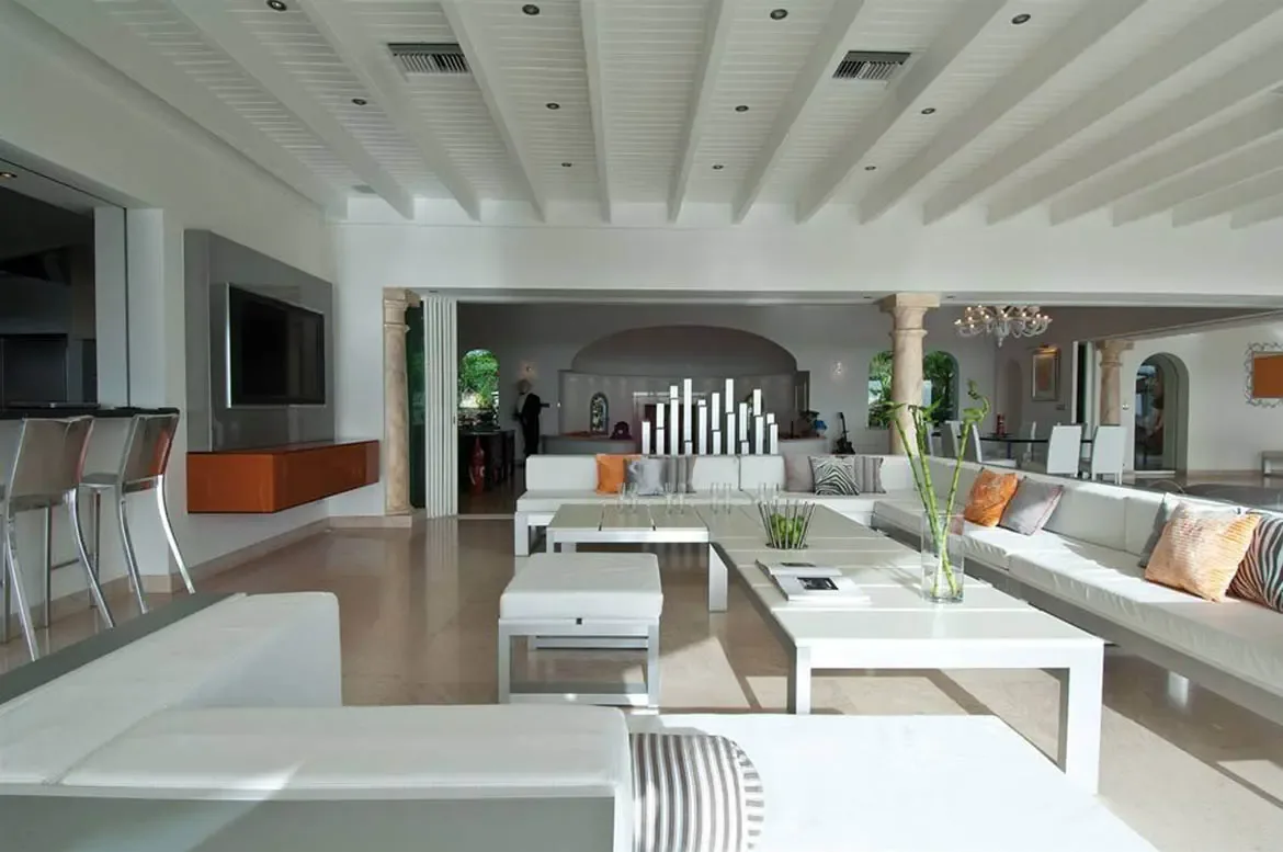 A white U-shaped sofa with zebra-print and orange pillows surrounds glass coffee tables with bamboo stalks, where a bar with chrome stools and a flat-screen TV anchor the vaulted space.