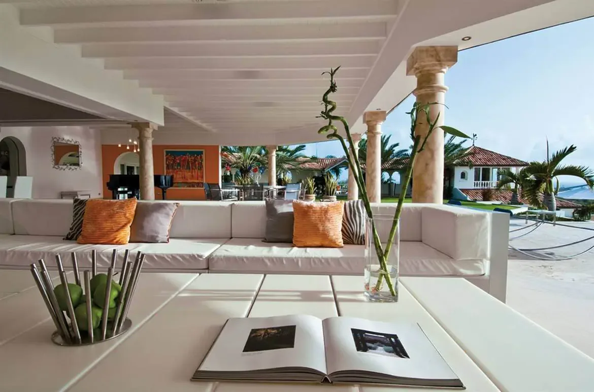 A white L-shaped sofa with zebra-print and orange pillows sits under stone columns, where a grand piano faces an orange accent wall and an outdoor terrace.