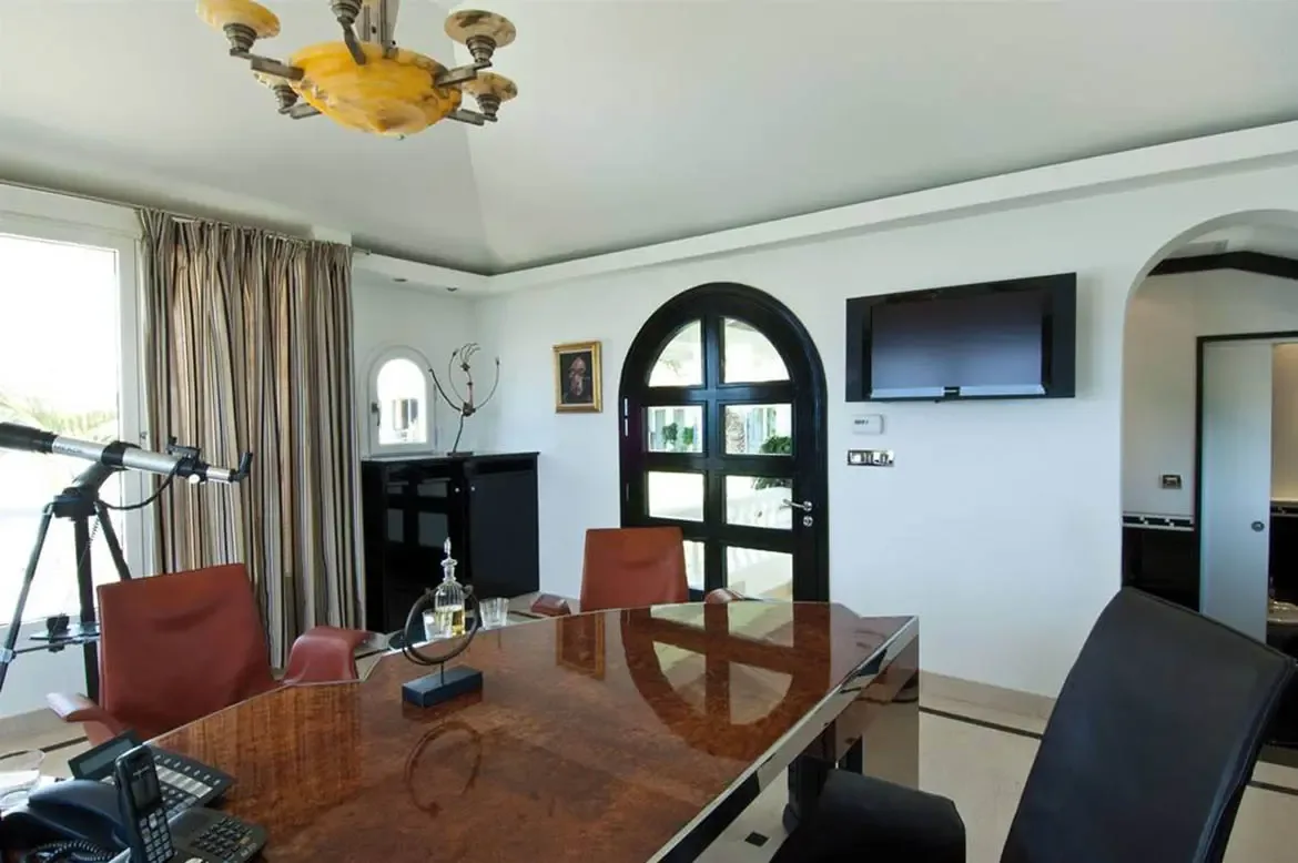 A polished burled-wood desk sits before striped curtains and an ocean-view window, where a telescope and an amber-glass chandelier fill a vaulted home office.