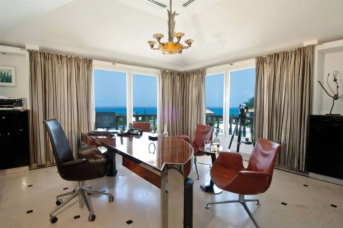 A curved chrome desk with a leather chair faces floor-to-ceiling windows framing a blue ocean, where cognac leather chairs and an Art Deco chandelier fill a marble-floored office.