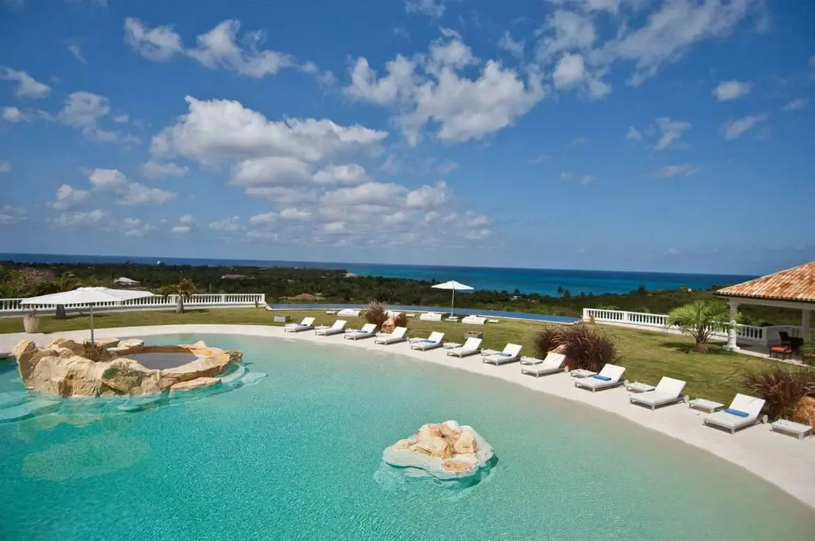 White loungers line a turquoise lagoon pool with a sandy beach entry and rock grotto, where a terracotta-roofed veranda and green palms overlook distant islands.