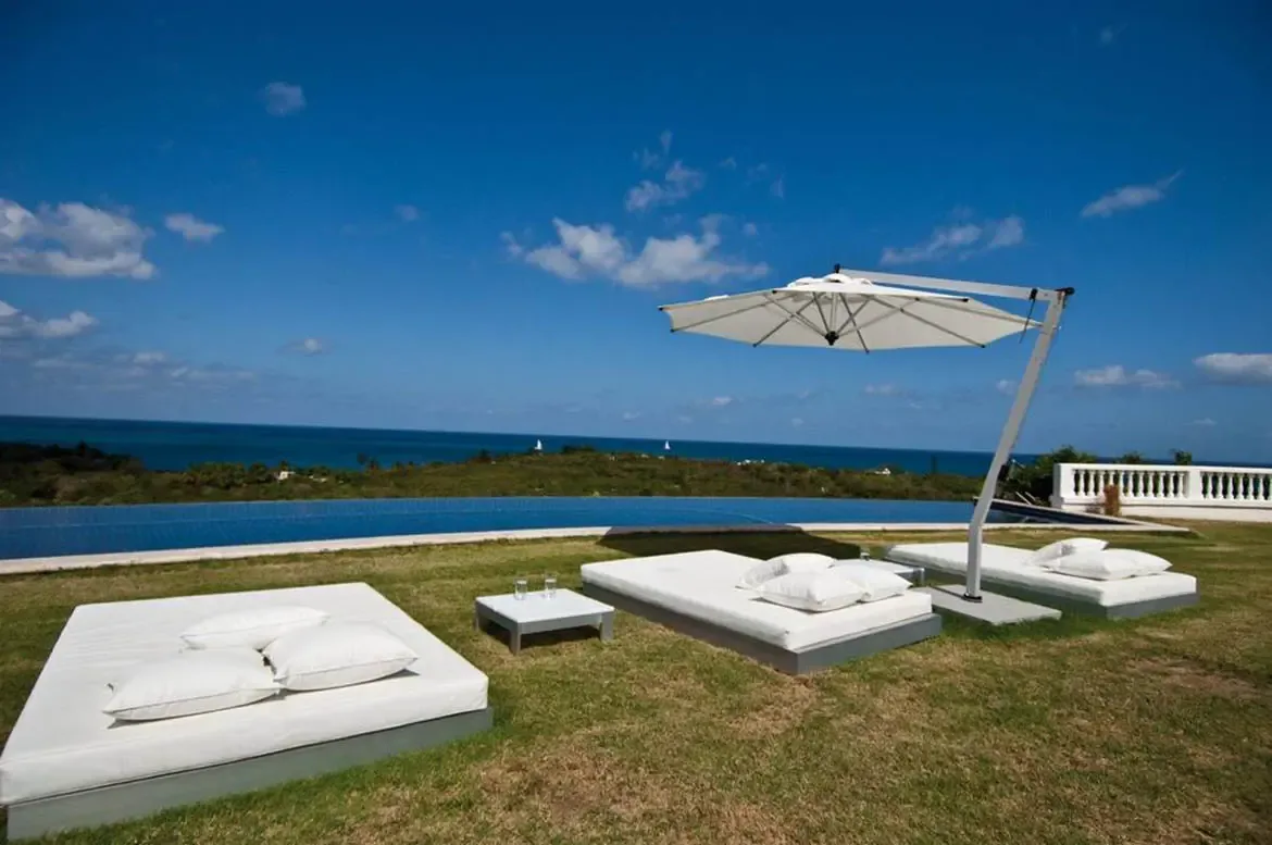 Three white daybeds with pillows sit on a green lawn under a cantilevered umbrella, where a narrow infinity pool merges with a deep blue ocean dotted with sailboats.