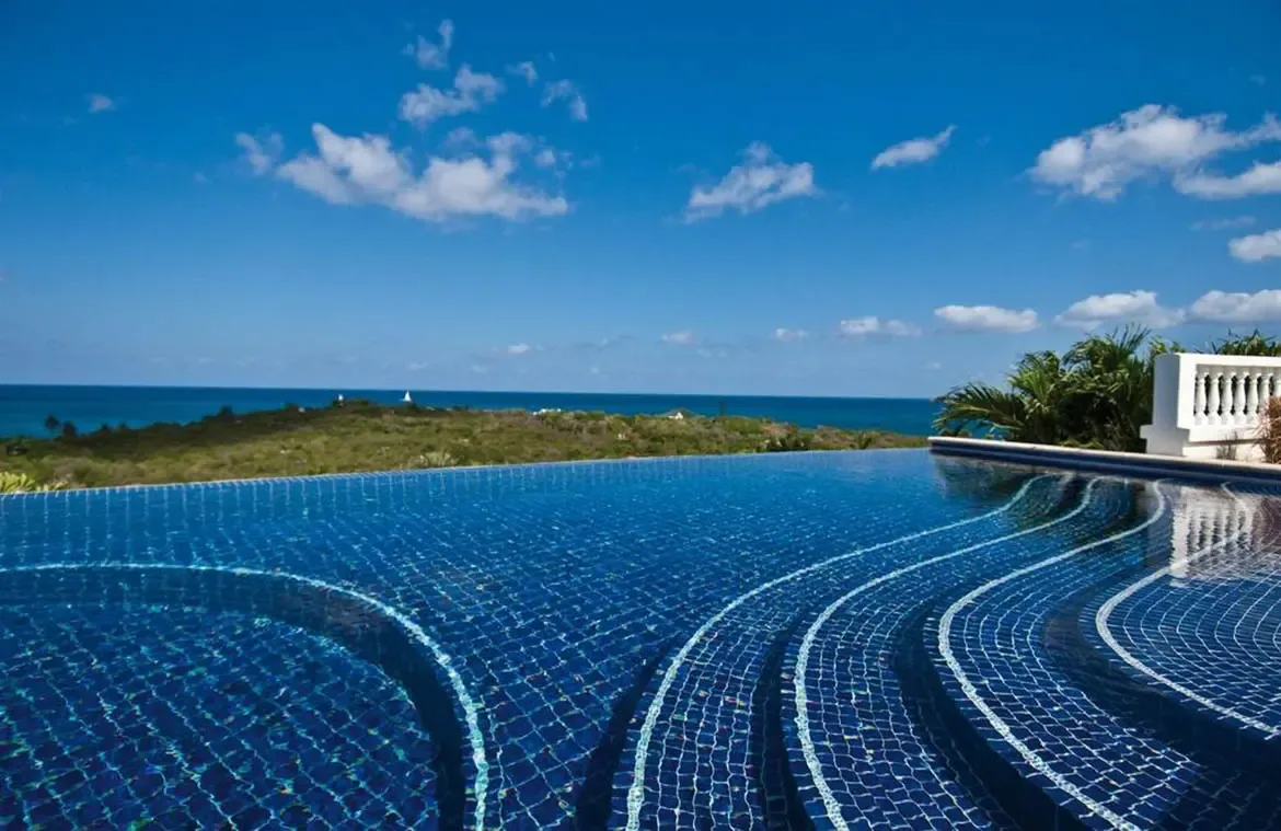 Deep blue mosaic tiles with white wave patterns line an infinity pool with submerged steps, where the edge merges with a turquoise ocean stretching to distant islands.