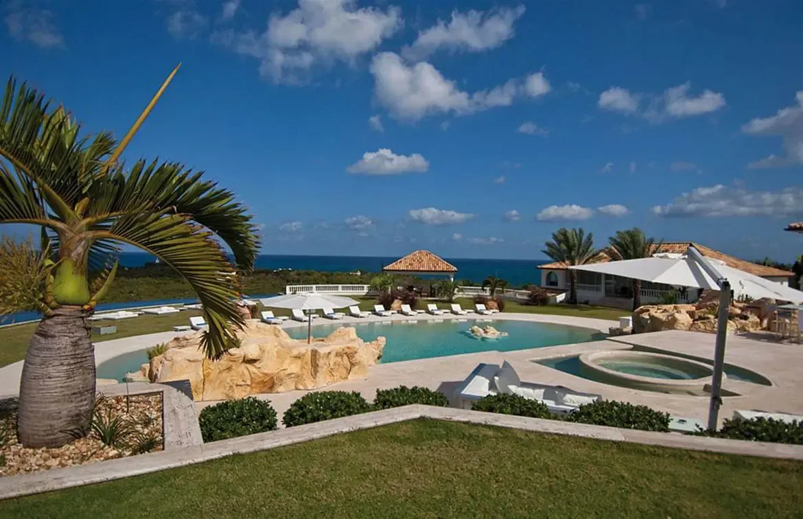 A turquoise lagoon-style pool with rock formations and beach entry curves beside a green lawn, where a palm tree and a circular hot tub overlook the deep blue ocean.