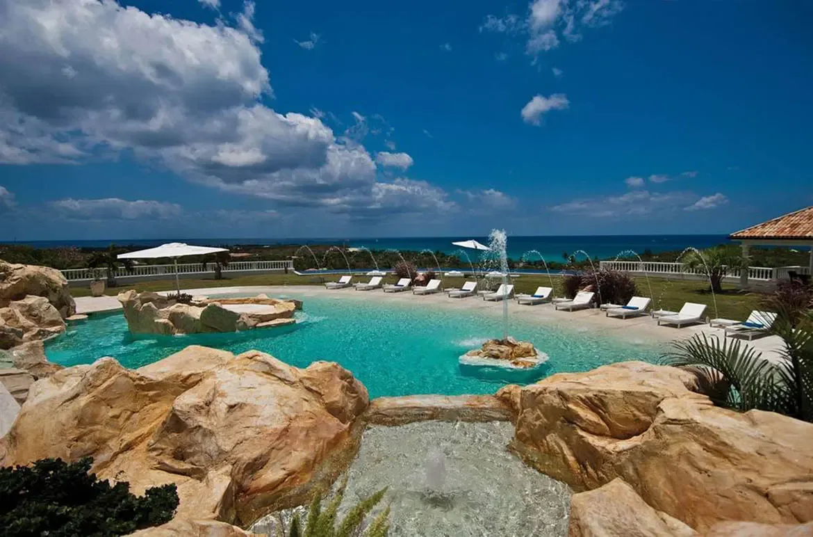 A turquoise lagoon-style pool with a rock fountain and beach entry sits beside white loungers on a green lawn, where water jets arch over a deep blue ocean panorama.