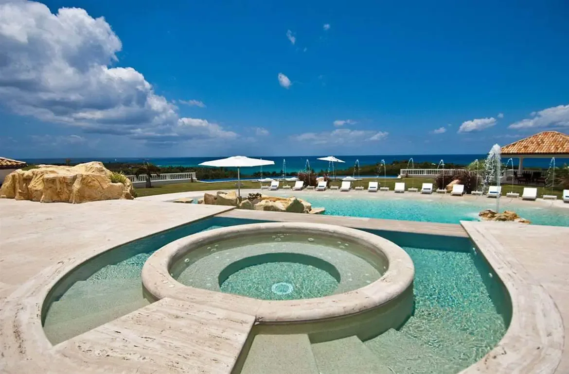 A tiered circular hot tub with stone steps descends into a turquoise infinity pool, where rock fountains arc water and white loungers overlook a deep blue ocean.
