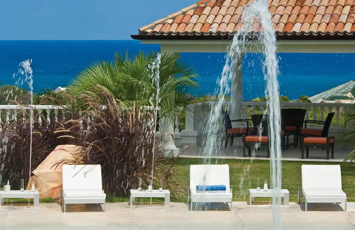 Water jets arc over a turquoise infinity pool beside white loungers and a green lawn, where a terracotta-roofed terrace with orange cushions overlooks a deep blue ocean.