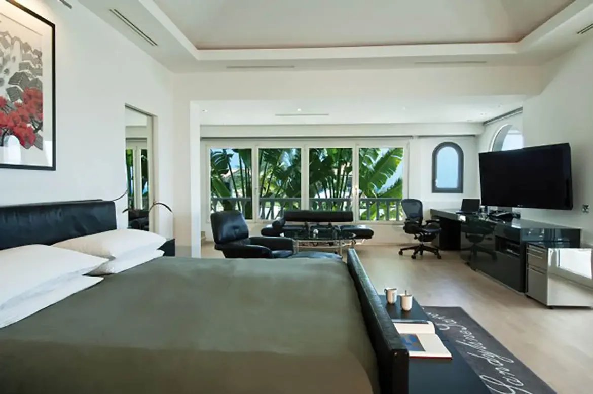 An olive-grey bed with a black bench sits before red flower art, where a black leather daybed and a workstation with twin monitors face floor-to-ceiling windows with palm trees.