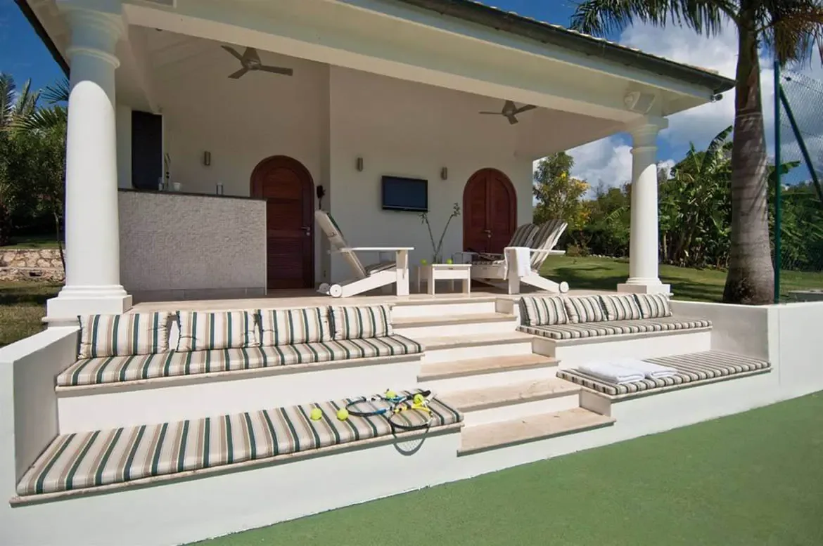 A white pavilion with columns houses a bar and flat-screen TV, where green-striped bench cushions and white loungers overlook a tennis court beneath a palm tree.