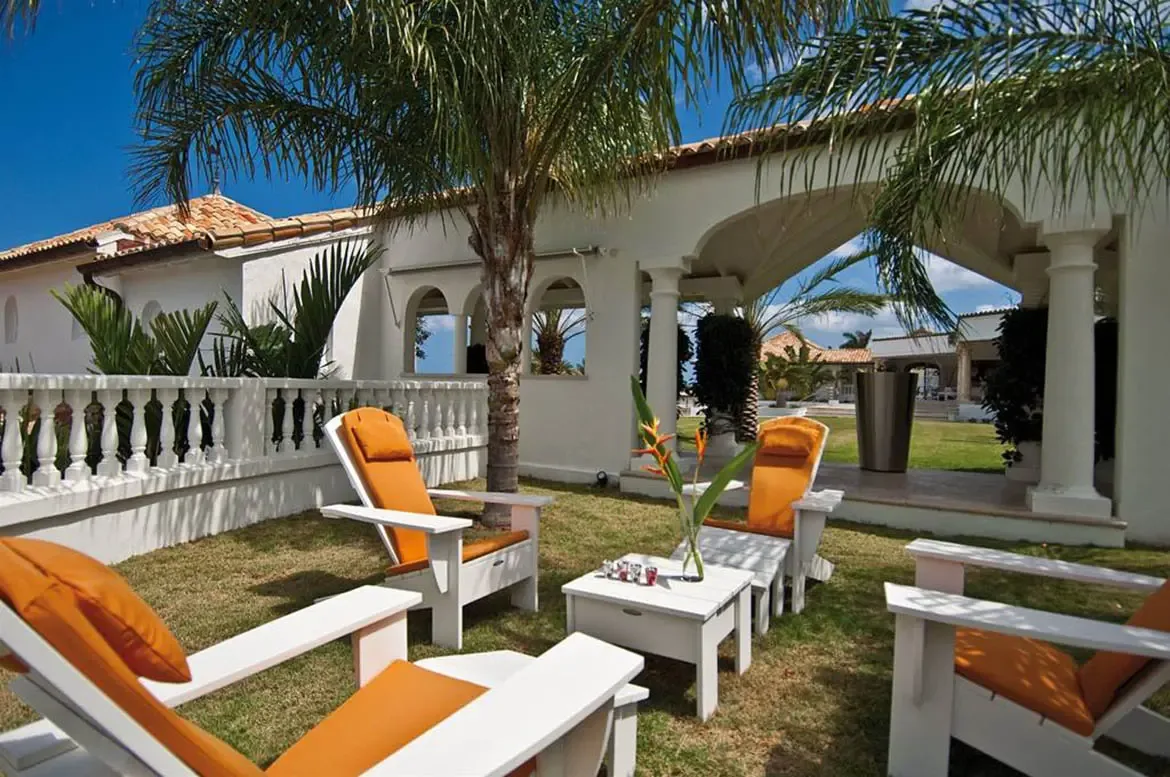 Orange-cushioned white loungers sit on a green lawn beside a white coffee table with a tropical flower, where a palm tree and arched white columns frame a terracotta-roofed veranda.