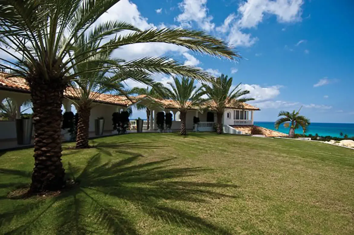 A white Mediterranean villa with terracotta roofs and arched columns stands beside palm trees on a green lawn, where the turquoise ocean stretches below a blue sky with white clouds.