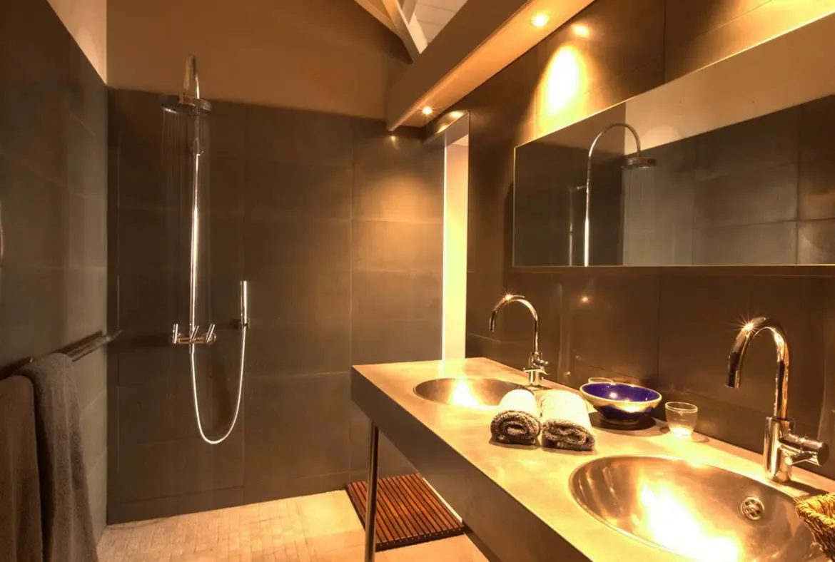 Two round metallic sinks sit on a dark vanity under a frameless mirror, where a walk-in rainfall shower with grey tiles and recessed lighting creates a spa-like atmosphere.