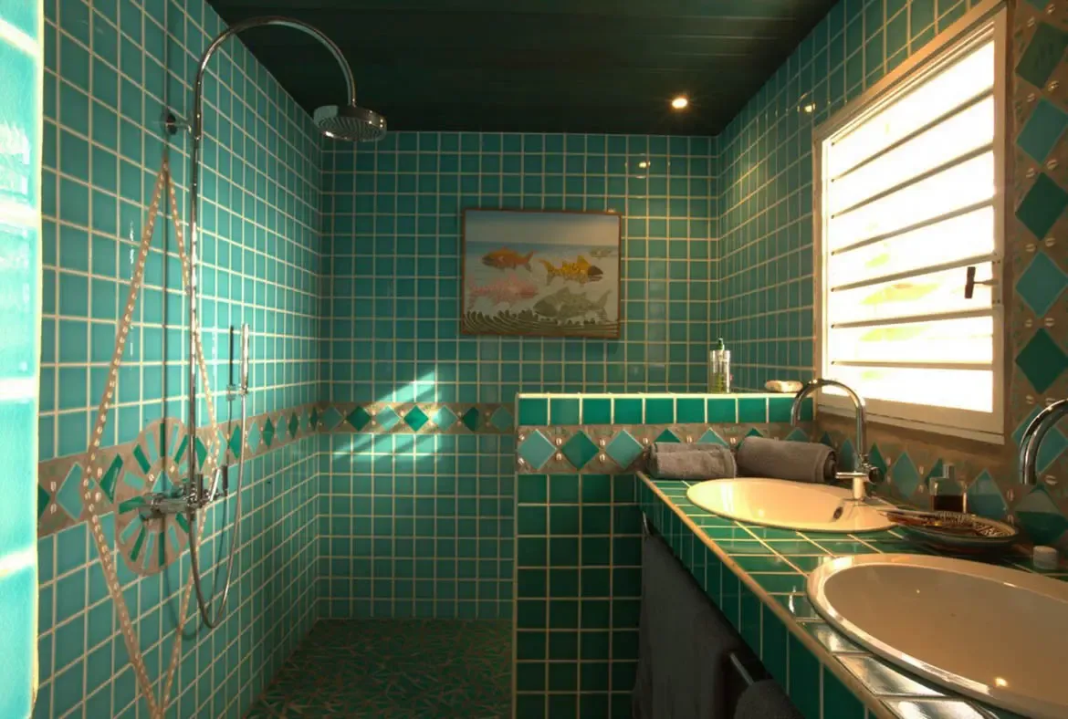 Wall-to-wall turquoise tiles with a diamond border wrap a double vanity with oval sinks, where a fish artwork and a curved rainfall shower complement the tropical design.