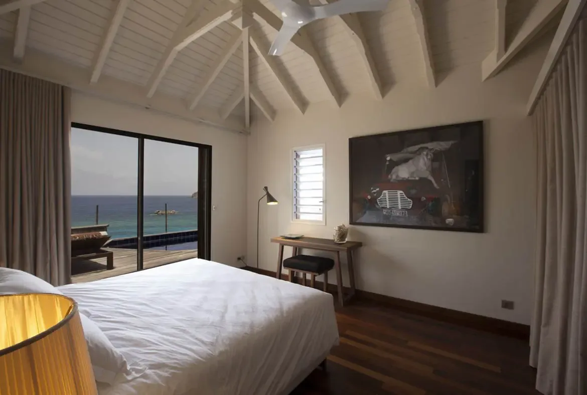 A white bed faces open doors where a pool deck meets turquoise ocean, while a framed photo of a vintage red car with a white dog and a gold-shaded lamp warm the vaulted bedroom.