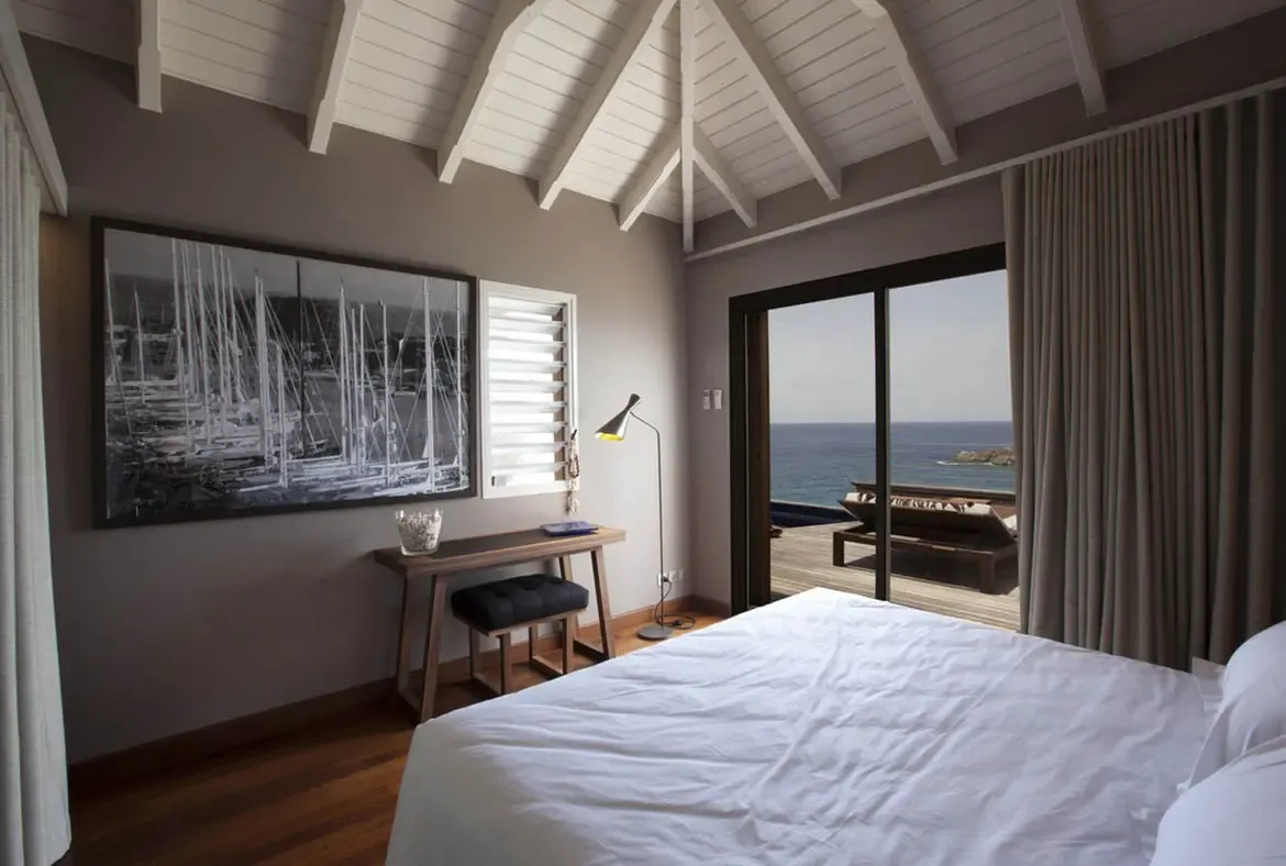 A white bed faces open-concept doors to a wooden deck and turquoise ocean, where a large sailboat photograph and a minimalist console add elegance under a vaulted ceiling.