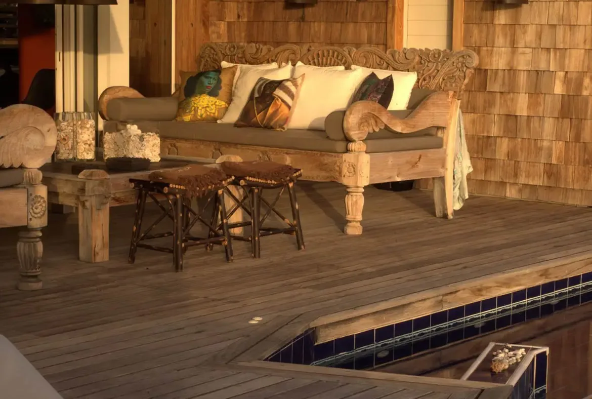 A weathered carved daybed with portrait pillows and taupe cushions sits beside branch-leg stools, where a blue pool edge and shingle wall catch the golden hour glow.
