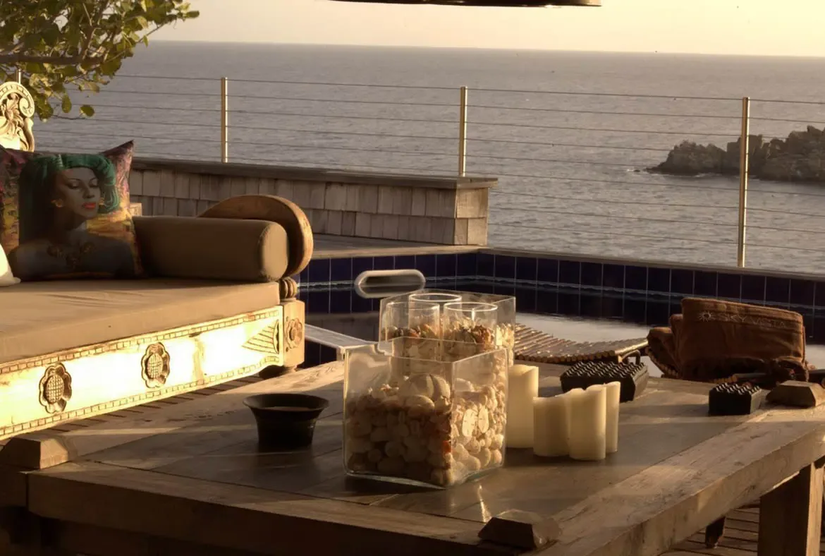 An ornate cream daybed with a green-haired portrait pillow sits beside a shell-filled glass vase and white candles, where a blue-tiled pool and ocean sunset warm the terrace.