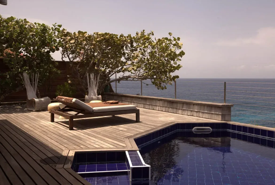 Wicker chairs with brown cushions tuck under leafy shade on a weathered deck, where a blue-tiled pool and wire railings open to deep ocean and distant islands.