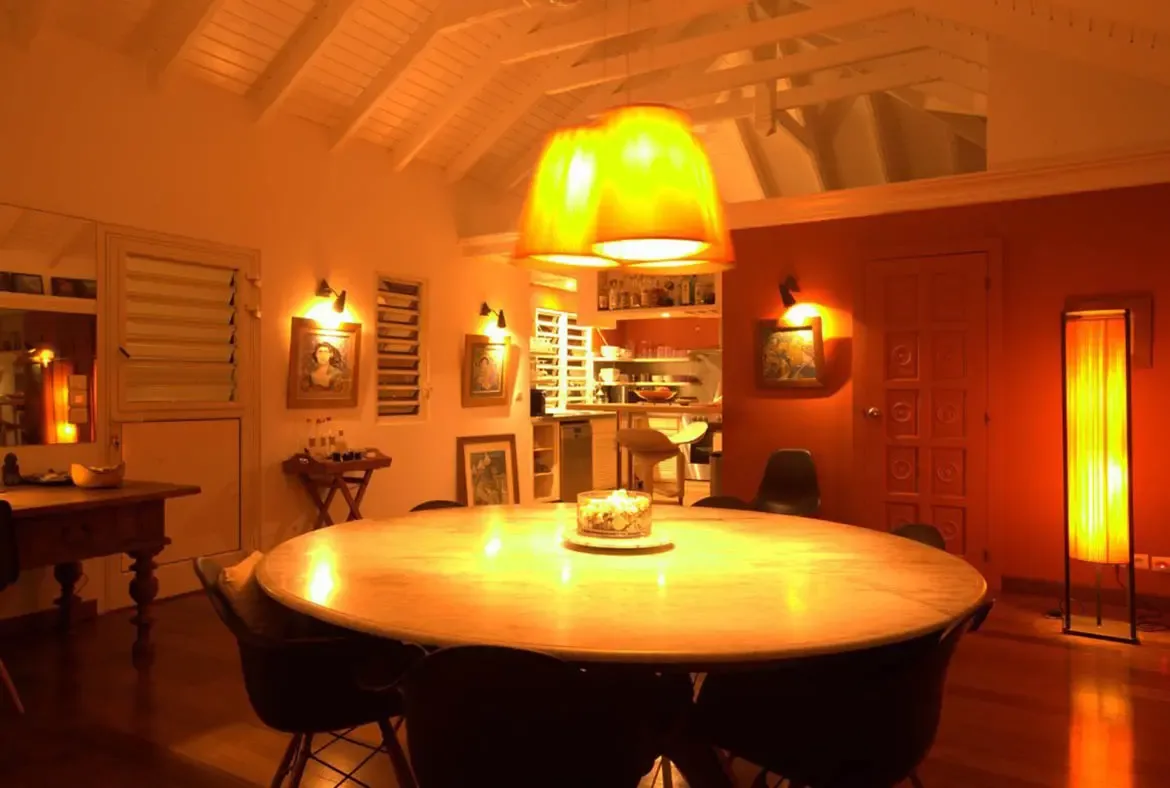 Golden light from orange pendants and spotlights illuminates a round marble table and a terracotta wall with art, where an open arch reveals a kitchen with wine bottles on shelves.
