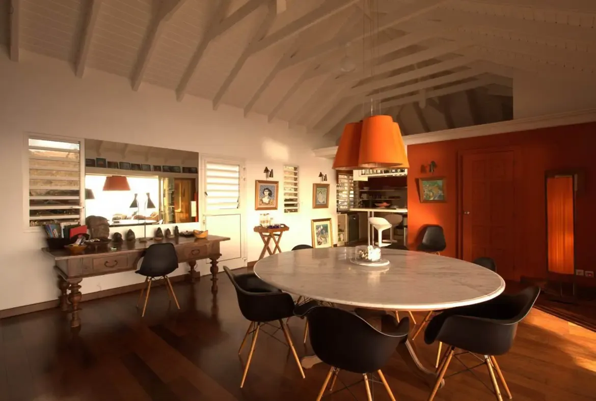 A round marble dining table with black chairs sits under a three-tier orange pendant light, where a terracotta accent wall and curated art collection warm a vaulted-ceiling space.