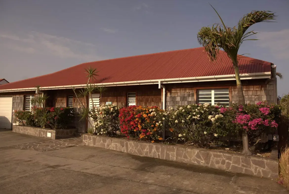 A red corrugated roof caps weathered shingle walls, where magenta and orange bougainvillea bloom behind a stone wall and a palm sways over a quiet courtyard.