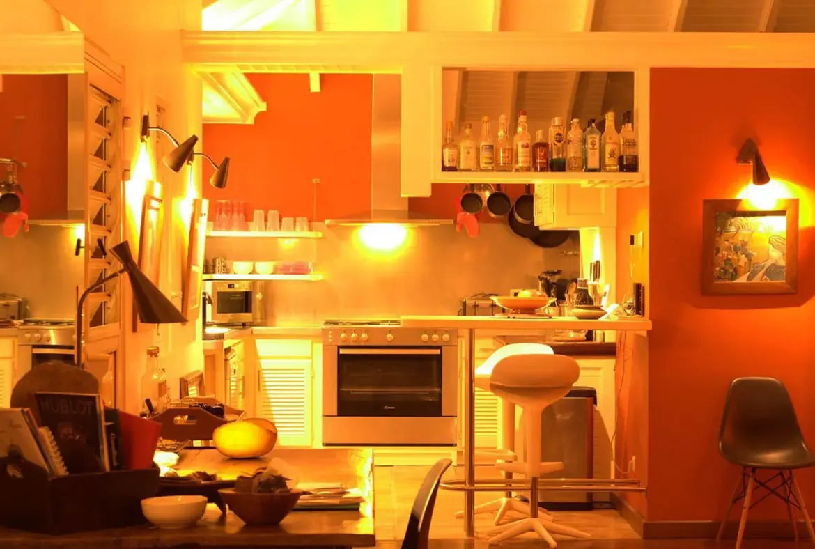 A compact kitchen with stainless steel stove and hanging pans glows warm orange, where a breakfast bar and a liquor cabinet stocked with bottles line a cozy dining nook.