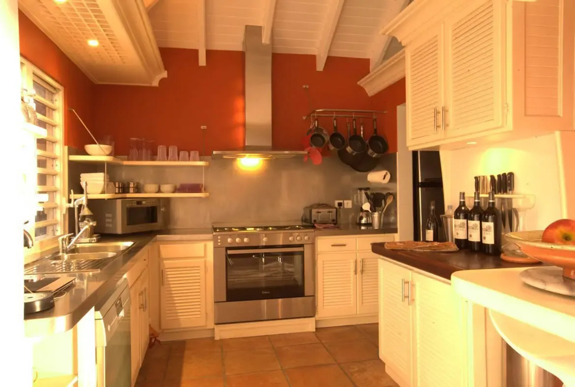 A professional stainless steel range sits between white louvered cabinets and an orange accent wall, where open shelves hold glassware and a baguette rests on a wooden island.
