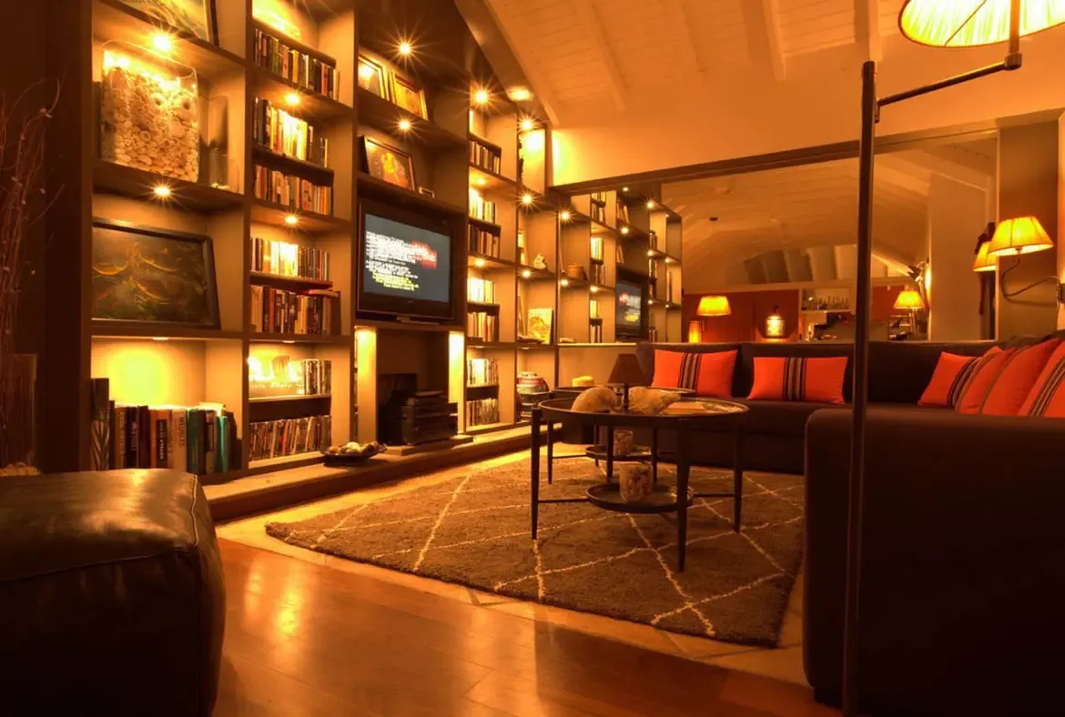 A floor-to-ceiling dark bookshelf with warm spotlights fills the wall beside a sofa with orange pillows, where glass-topped tables and a diamond-pattern rug warm the vaulted room.