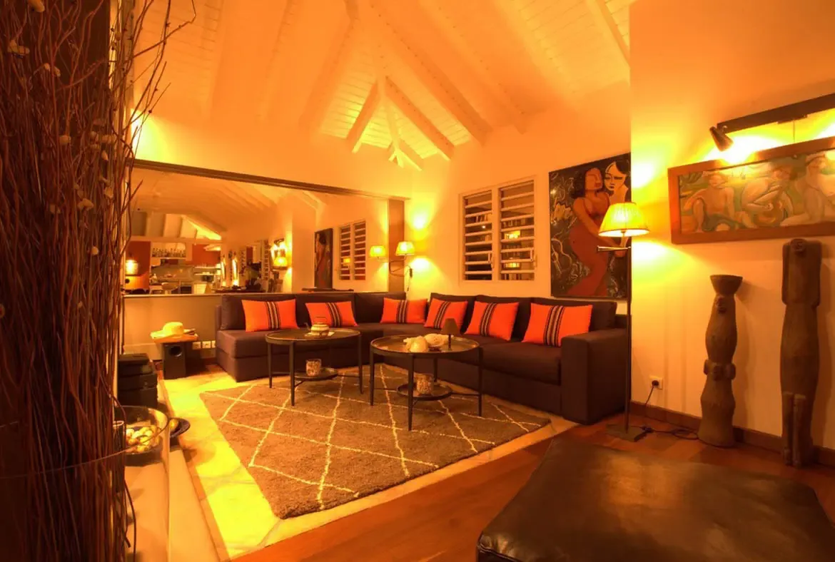 A dark L-shaped sofa with orange striped pillows sits under a vaulted ceiling, where tribal sculptures and warm amber lighting create a cozy eclectic living room.