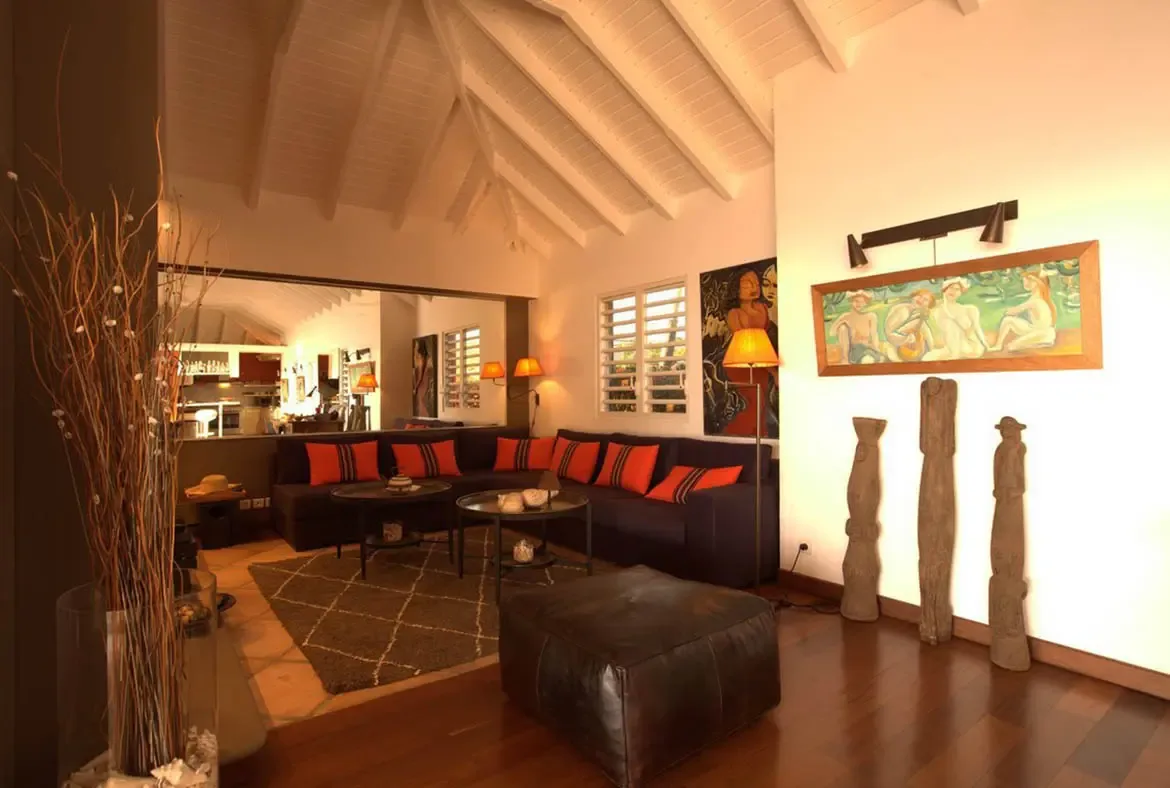A dark L-shaped sectional with orange and red pillows sits under a vaulted ceiling, where tall tribal totems and a horizontal painting anchor the warm eclectic living room.