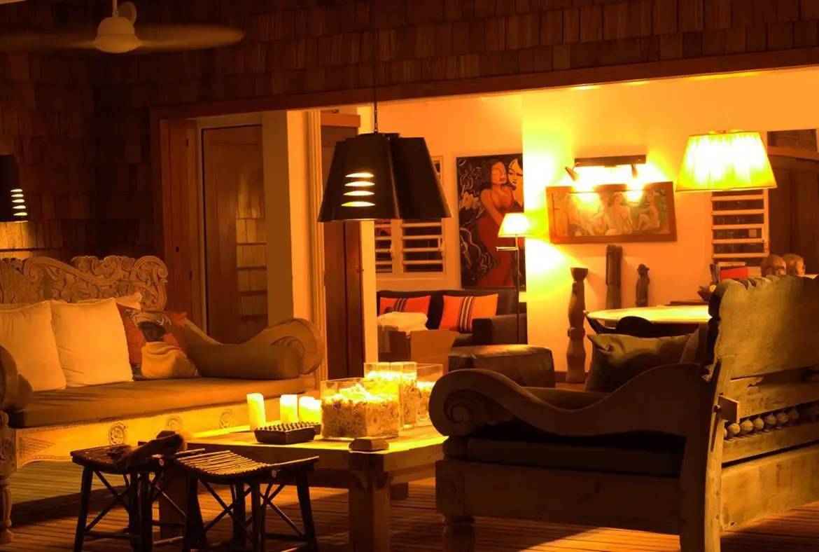 At night, carved wooden sofas with white cushions glow under cone pendant lights, where candles in glass jars and warm amber light transform the terrace into a cozy retreat.