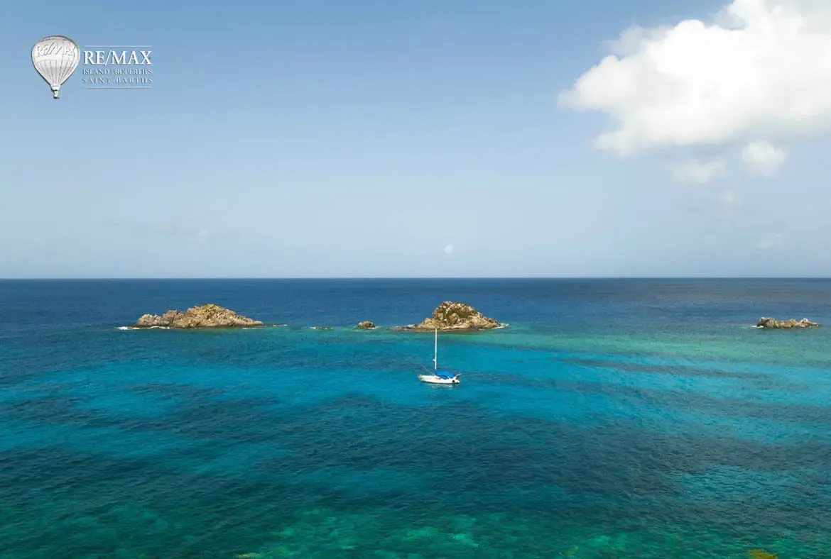 A sweeping ocean panorama reveals turquoise shallows around rocky islets and a lone white sailboat, where deep blue water meets a pale sky on the horizon.
