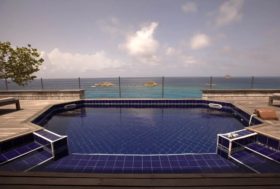 A cobalt-blue tiled pool with steps reflects the sky on a wooden deck, where glass railings and swaying palms frame turquoise ocean dotted with rocky islands.