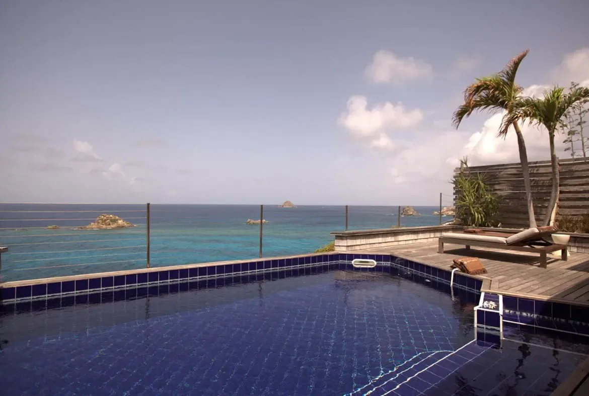 A white cushioned lounger sits on a weathered wood deck beside a blue-tiled pool, where wire railings and leaning palms frame turquoise ocean and rocky islets.