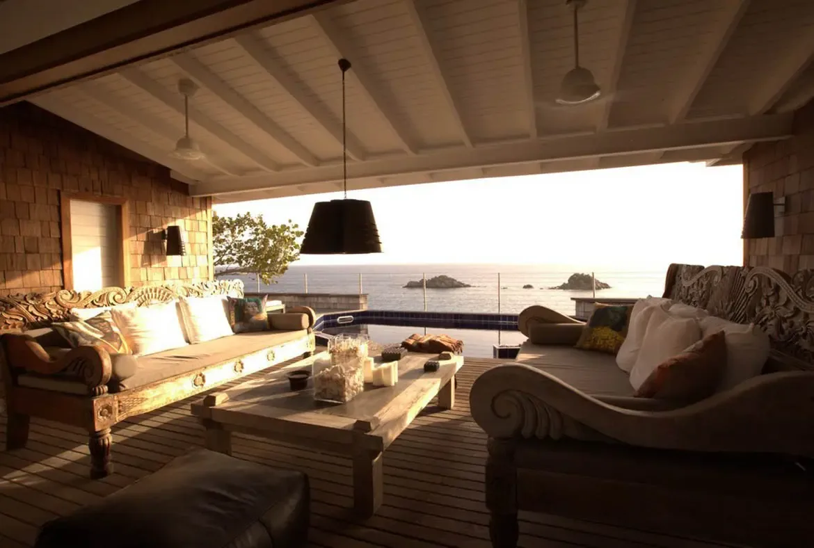 Two carved wooden daybeds with white cushions face each other across a shell-filled coffee table, where a plunge pool meets glass railings and a vast ocean beyond.