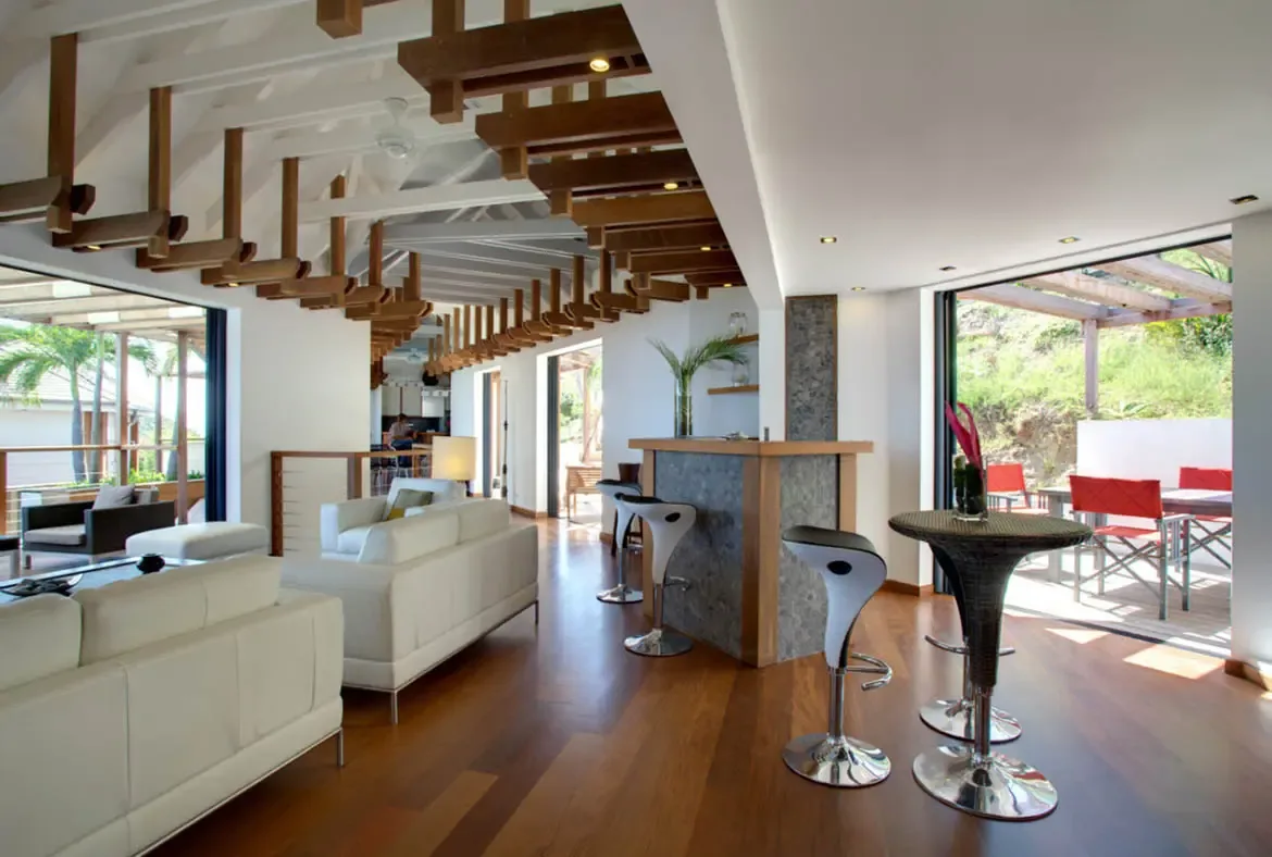 A stone-clad bar with white stools and a glass dining table sit under a latticed ceiling, where open doors lead to a shaded pergola with red dining chairs overlooking lush hillside greenery.