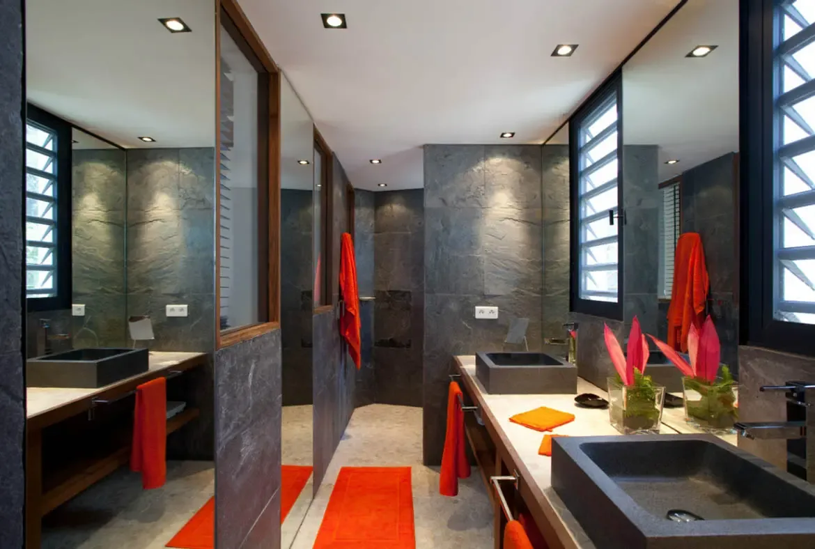 Dark slate walls and minimalist stone vessel sinks anchor a bold bathroom, where red towels, orange bromeliads, and recessed spotlights create dramatic contrast against frameless mirrors.