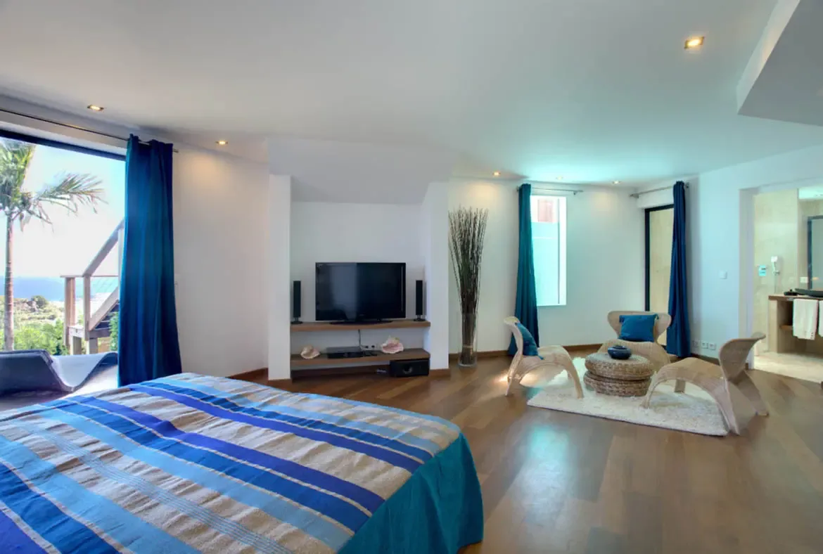 A bed with blue-striped bedding sits before open glass doors framed by navy curtains, where a TV niche, wicker chairs, and a deck overlooking turquoise ocean complete the suite.